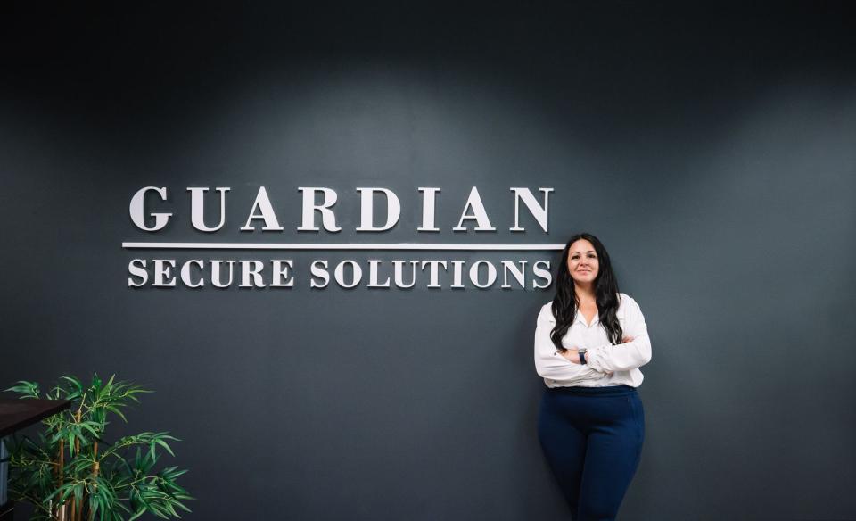 A woman stands with arms crossed in front of a dark wall displaying the words "Guardian Secure Solutions.