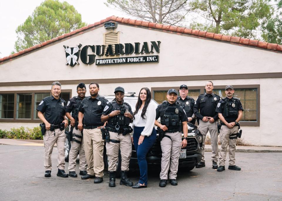 A group of security personnel in uniform stands in front of a building labeled "Guardian Protection Force, Inc." with a woman in business attire.