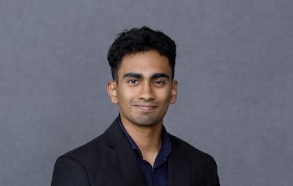 A man wearing a black suit and dark shirt stands against a plain gray background, looking at the camera with a neutral expression.