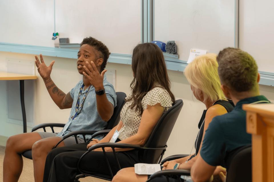 Four people sit in chairs in a classroom setting; one person gestures with both hands while speaking, and the others listen attentively.