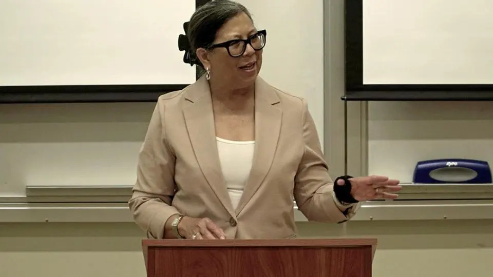 A woman in a tan blazer and glasses speaks at a wooden podium in a classroom setting, gesturing with one hand.