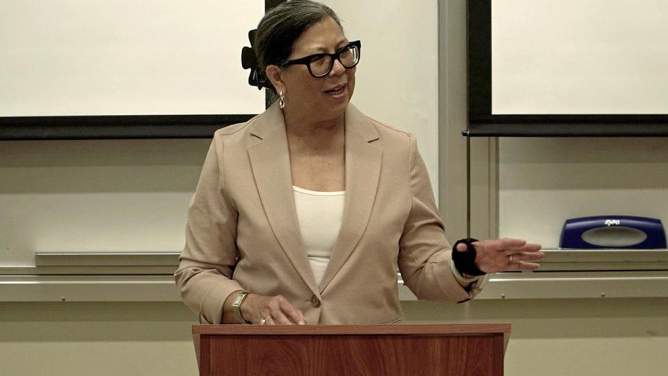 A woman in a tan blazer and glasses speaks at a wooden podium in a classroom setting, gesturing with one hand.