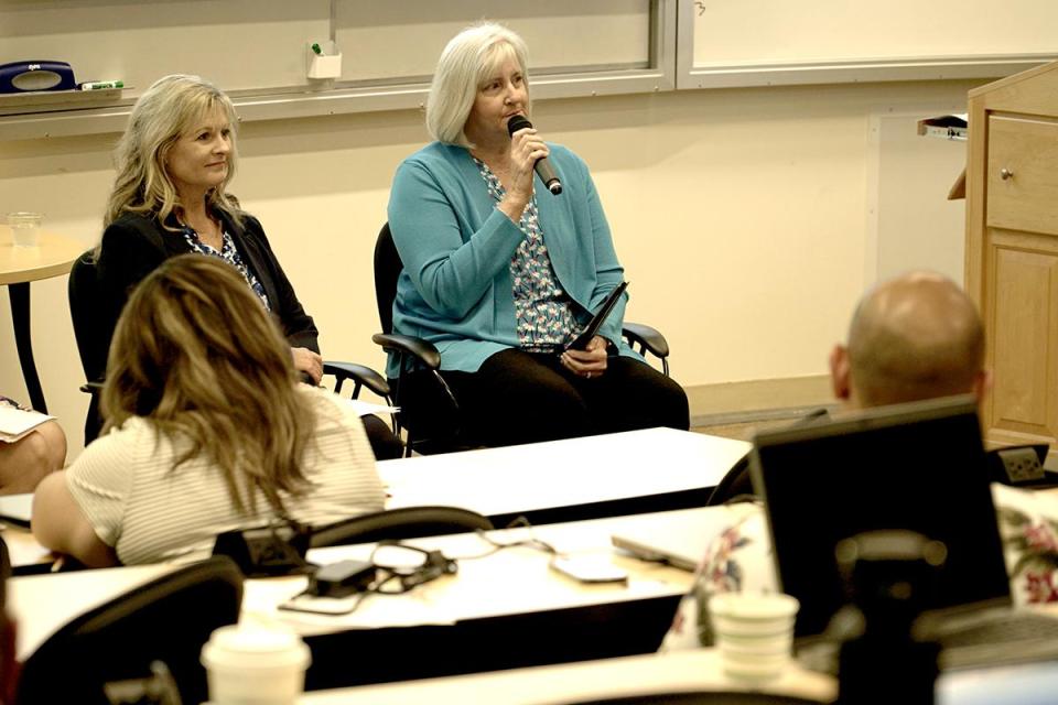 Two women sit at the front of a classroom; one speaks into a microphone while the other listens. Several people are seated facing them.