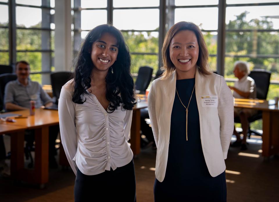 Rachel Johnson and Ayung Tseng together in Executive Board Room with people seated at a table in the background.