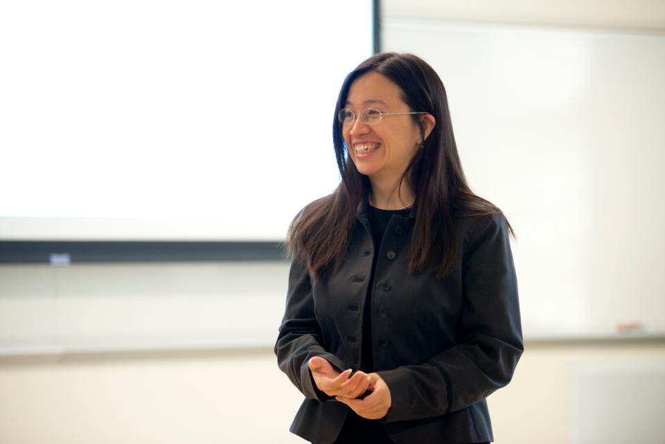 Professor Rachel Chen with long dark hair, wearing glasses and a black jacket, stands and smiles in a well-lit classroom or lecture room.