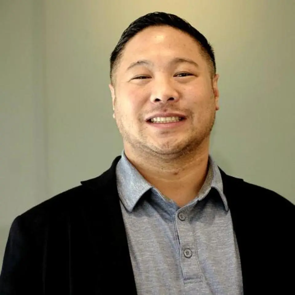 A man with short dark hair smiles at the camera, wearing a light gray collared shirt and a black blazer against a neutral background.