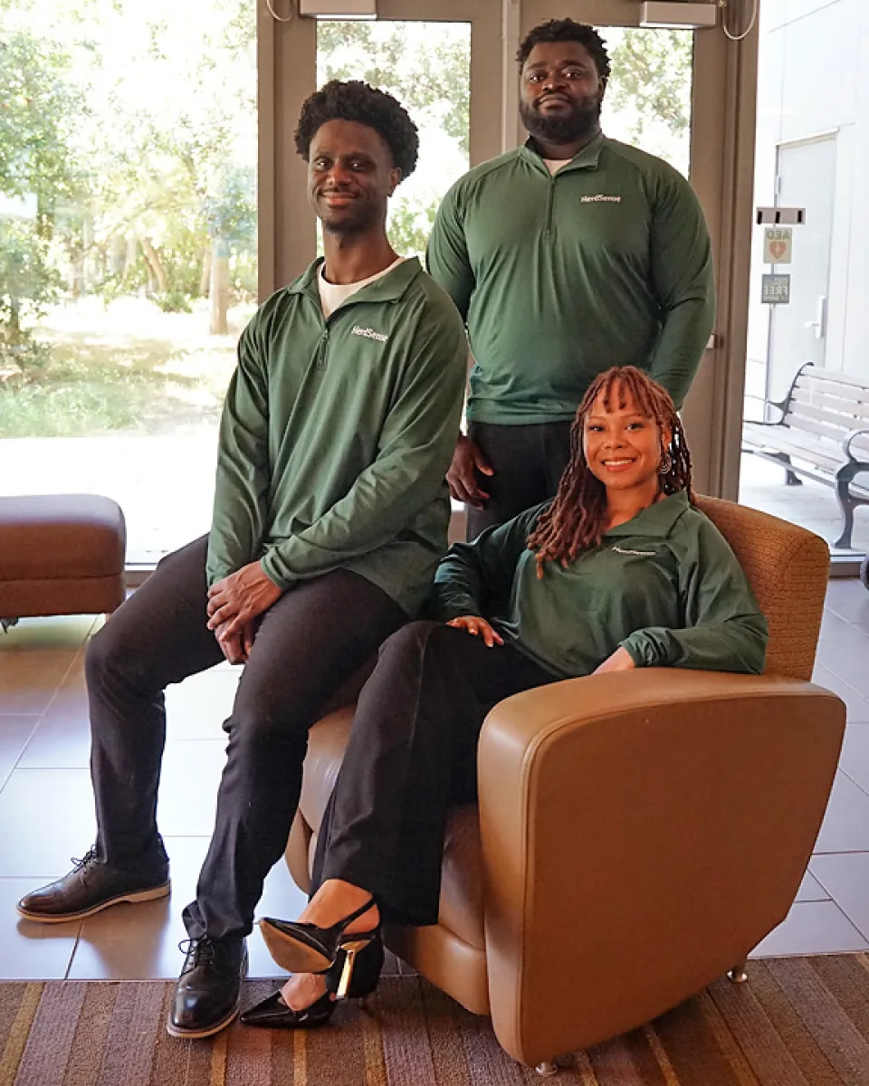 Three people wearing matching green long-sleeve shirts pose indoors; two are standing, and one is seated in a brown chair. Trees and a bench are visible outside through large windows.