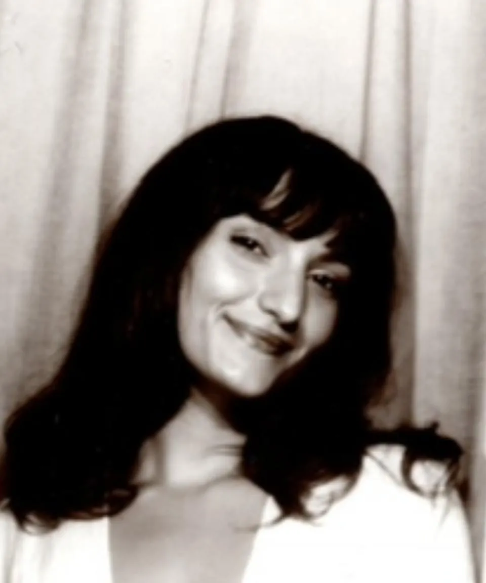 A woman with medium-length dark hair and bangs smiles at the camera in a black-and-white portrait with a curtain backdrop.