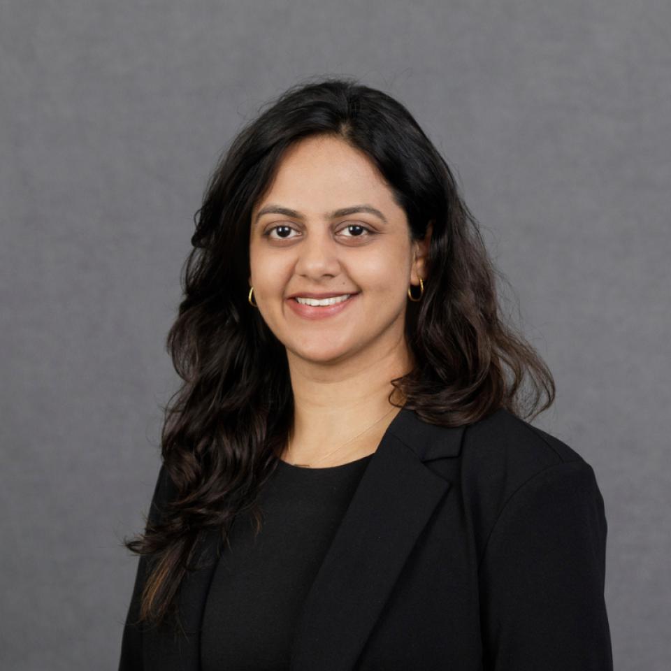 Woman with long dark hair, wearing a black blazer and top, smiling at the camera against a plain gray background.