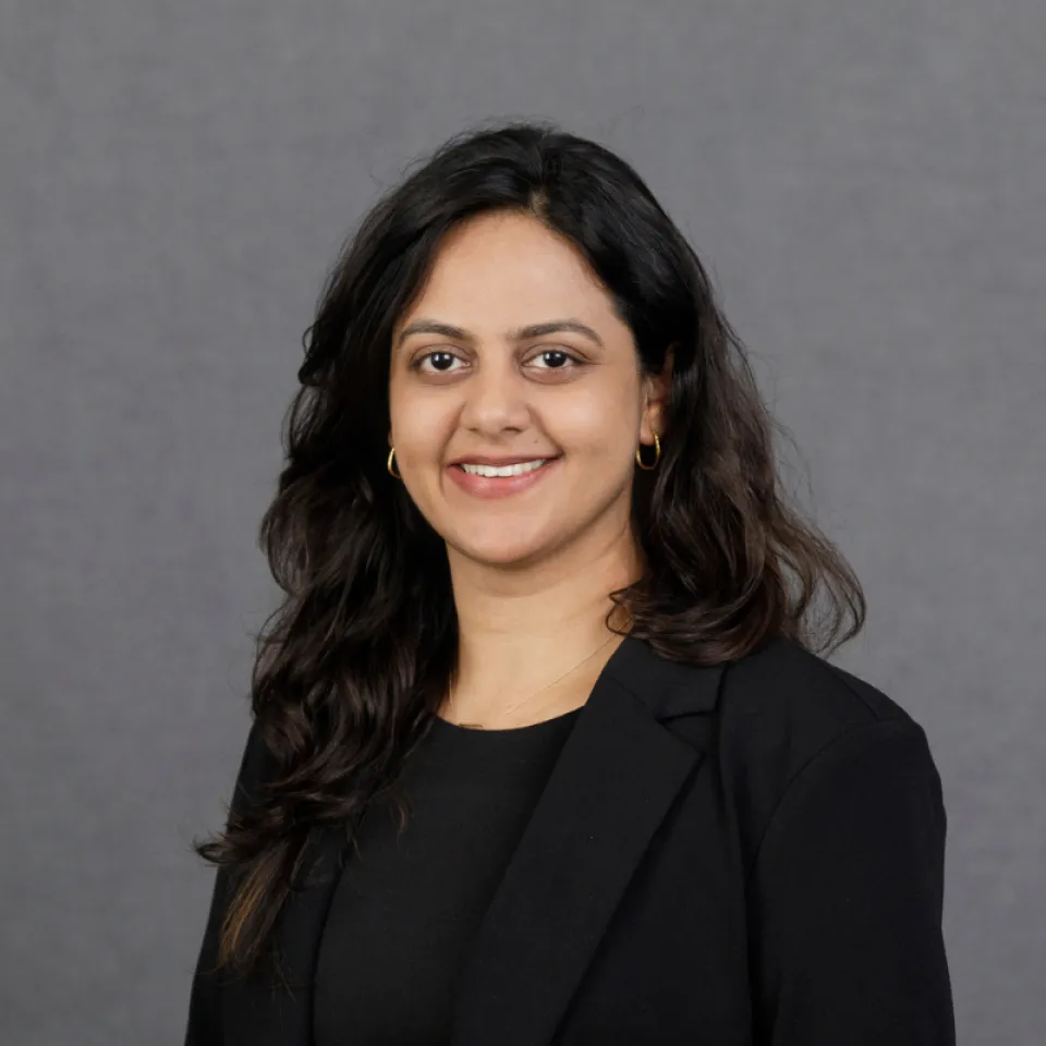 Woman with long dark hair, wearing a black blazer and top, smiling at the camera against a plain gray background.