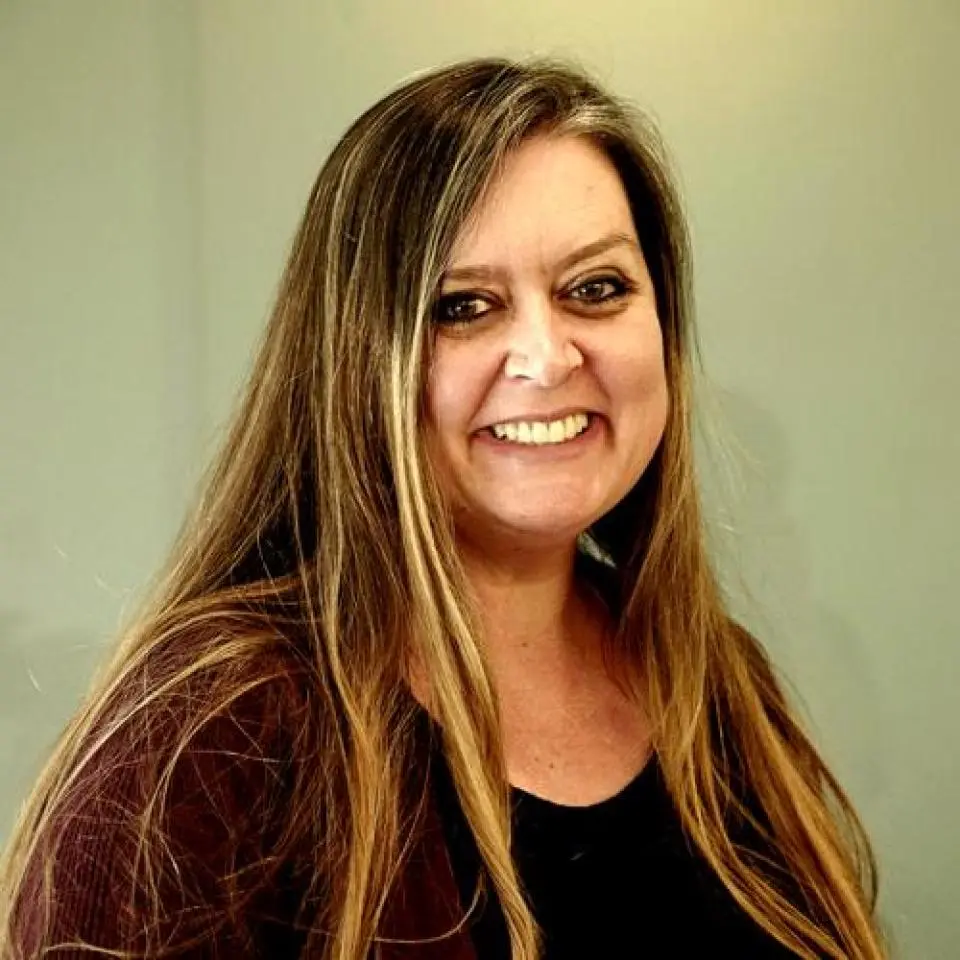 Woman with long, straight brown hair wearing a dark top and smiling, standing in front of a plain, light-colored background.