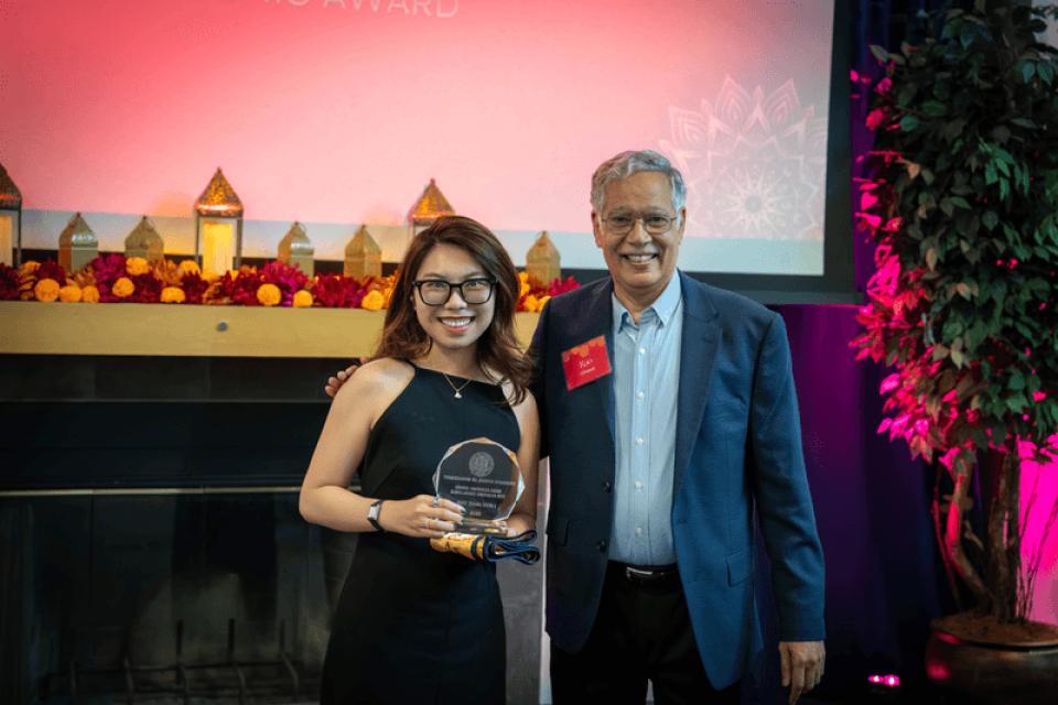 Two people stand side by side smiling; the woman on the left holds an award plaque. They are at an indoor event with a decorated table and a projection screen in the background.