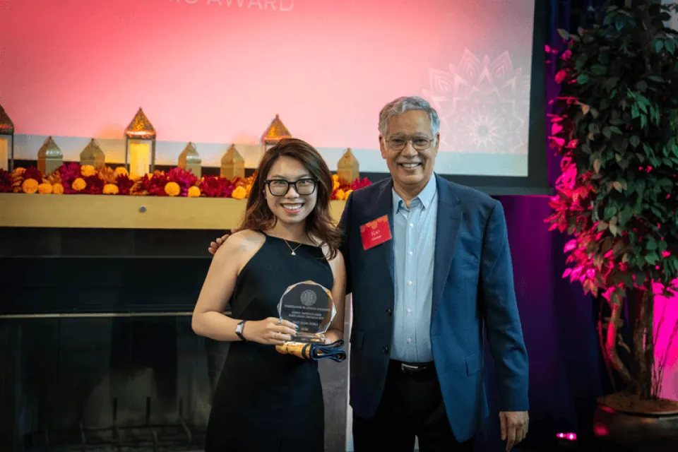 Two people stand side by side smiling; the woman on the left holds an award plaque. They are at an indoor event with a decorated table and a projection screen in the background.