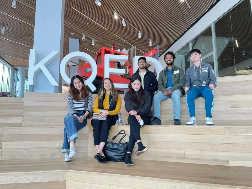 Six MSBA team members sitting on wooden steps inside KQED modern building with large "KOE" letters and a red structure in the background.