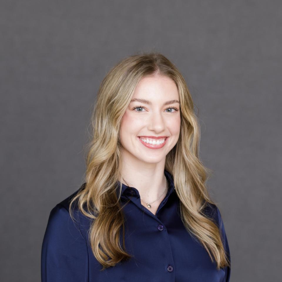 A young woman with long, wavy blonde hair wearing a dark blue shirt, smiling in front of a plain gray background.