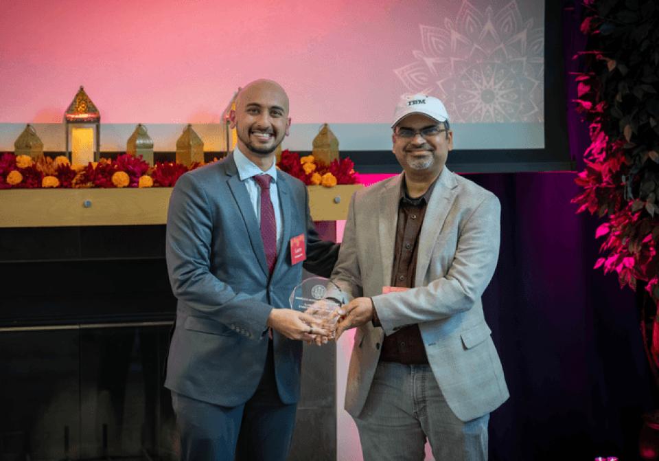 Two men stand side by side; one hands an award plaque to the other. Both are smiling and wearing suits. A decorated shelf and a projector screen are in the background.