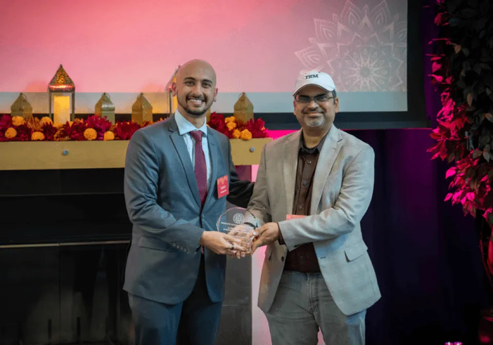 Two men stand side by side; one hands an award plaque to the other. Both are smiling and wearing suits. A decorated shelf and a projector screen are in the background.