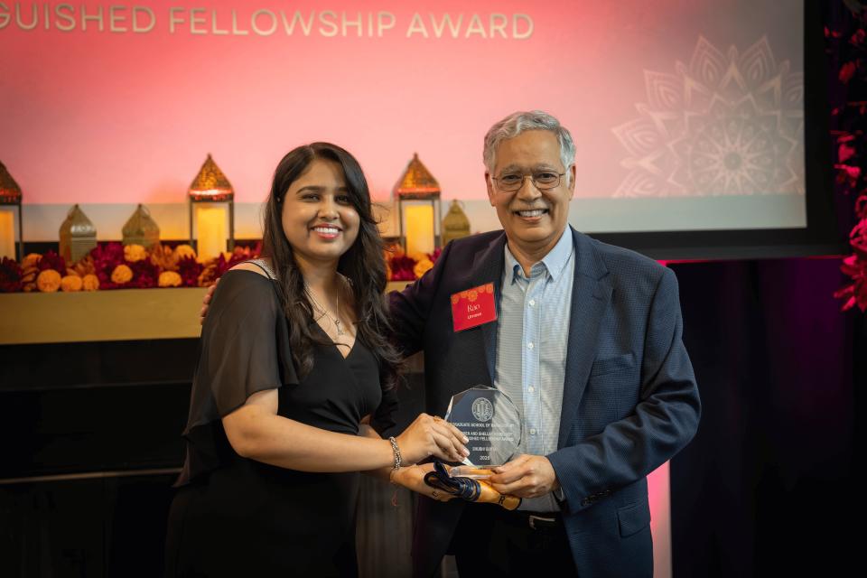 Two people stand together smiling as one presents an award to the other in front of a decorated stage with a "Distinguished Fellowship Award" sign.