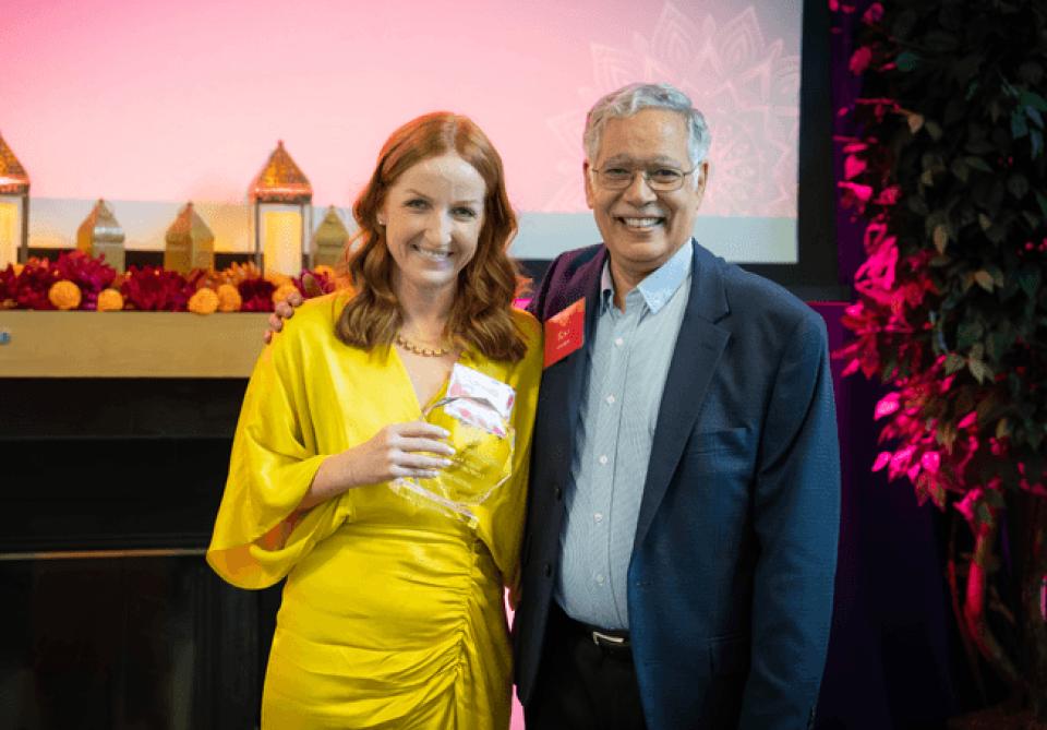 A woman in a yellow dress holding an award stands next to a smiling man in a suit at an indoor event with festive decor.