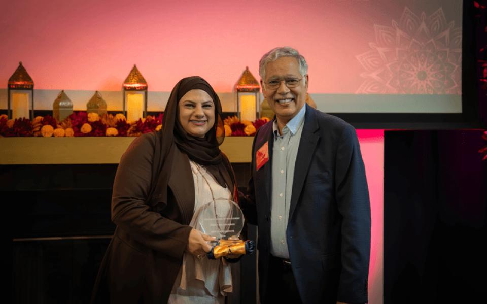 Two people stand side by side at an award ceremony, with one holding a glass trophy. Decorative lanterns and a patterned screen are in the background.