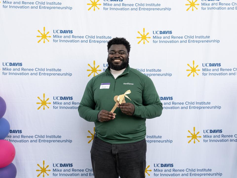 Jeremy Twumasi Ampofo in a green polo shirt holds a wooden award in front of a UC Davis backdrop.