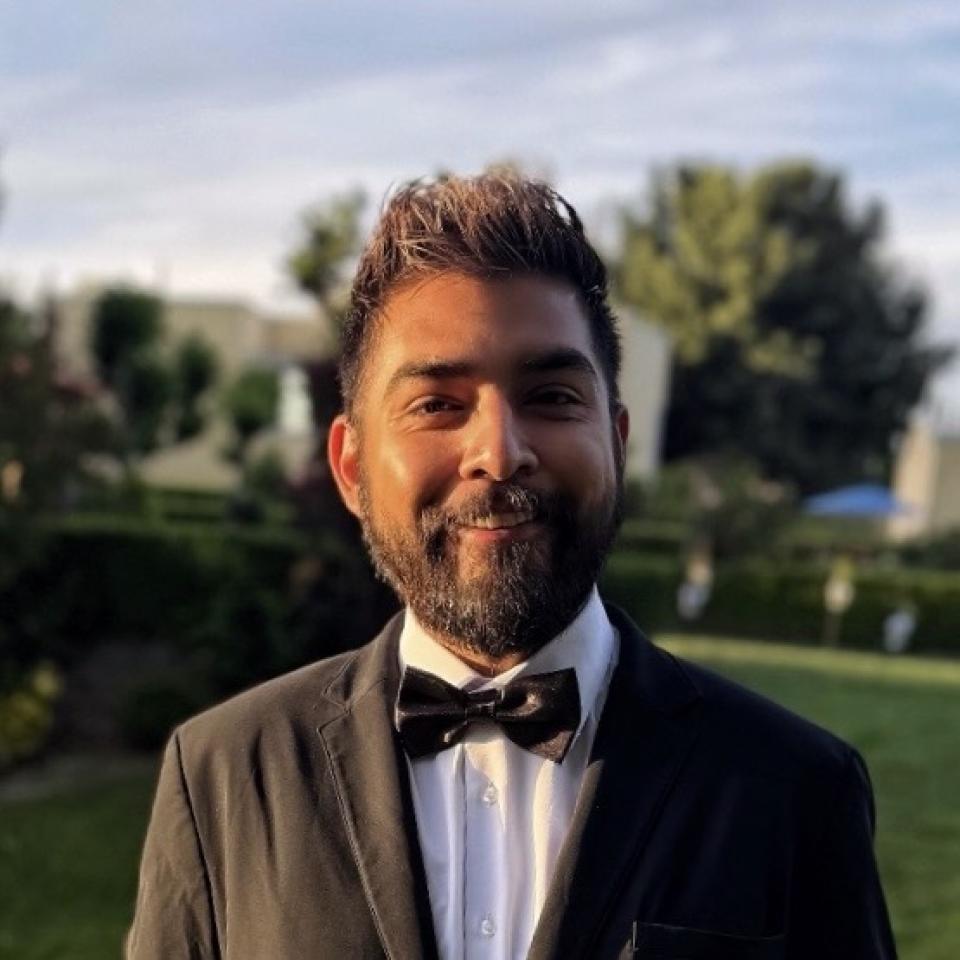 A man wearing a black tuxedo and bow tie stands outdoors in a garden setting, smiling at the camera with trees and greenery in the background.
