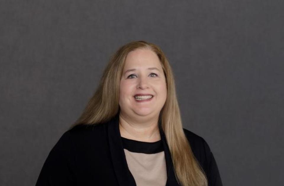 A woman with long blonde hair, wearing a beige top and black cardigan, stands smiling in front of a plain gray background.