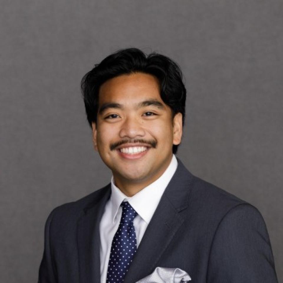 A man in a dark suit, white shirt, and polka dot tie smiles in front of a plain gray background.