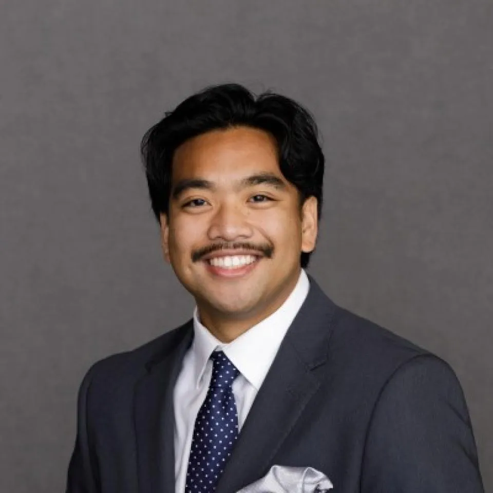 A man in a dark suit, white shirt, and polka dot tie smiles in front of a plain gray background.