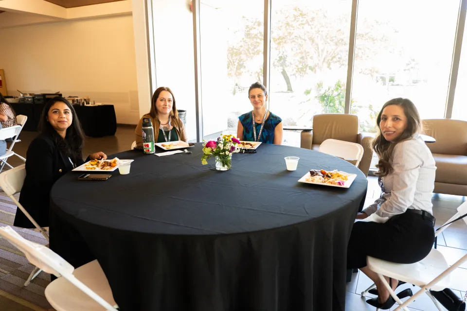 Four women seated around a round table with black tablecloth, eating lunch. Plates of food and drinks are on the table. The setting appears to be indoors with large windows in the background.