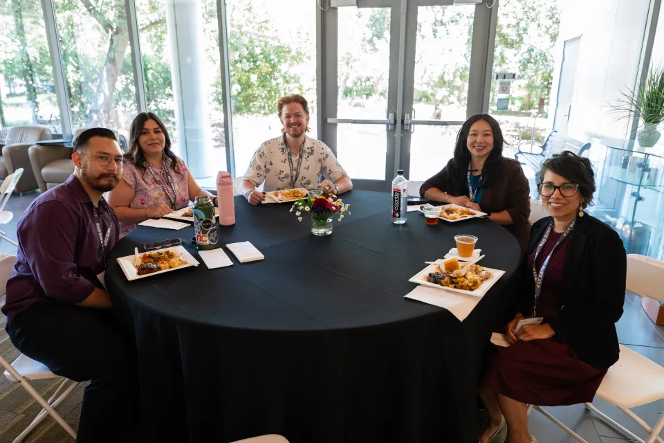 Five people sit around a round table with plates of food and drinks, smiling at the camera in a bright room with large windows and potted plants in the background.