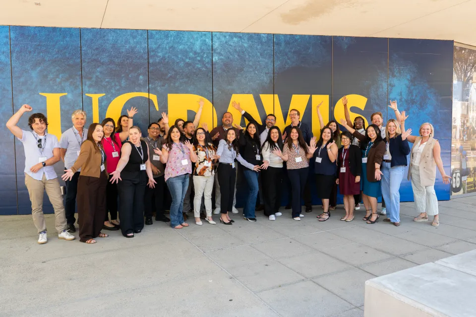 A group of people pose and smile in front of a blue and gold UC Davis sign outside a building on a sunny day.
