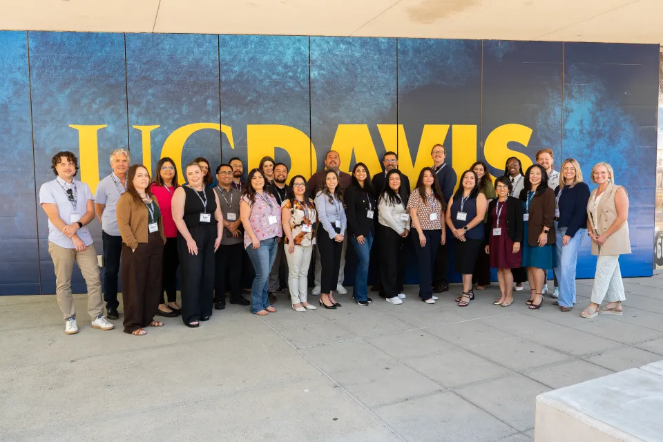 A group of people stands in front of a large UC Davis sign on a blue and yellow background, posing for a group photo outdoors.