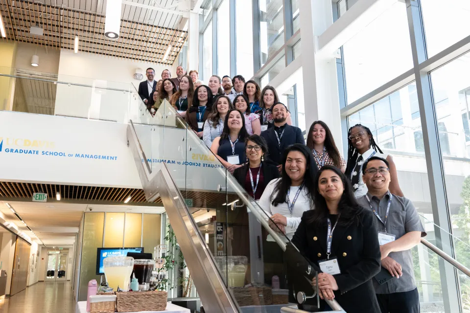 A group of people stand together on a staircase inside a modern building, posing for a group photo at the UC Davis Graduate School of Management.