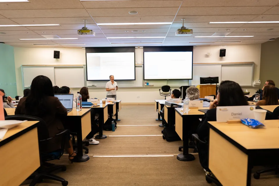 A person stands at the front of a classroom giving a presentation to seated students, with slides displayed on screens behind them.