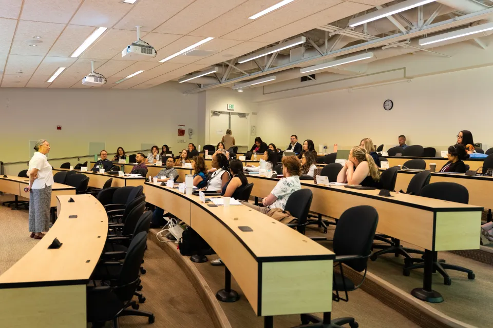 A woman stands at the front of a classroom speaking to students who are seated at curved desks, listening and taking notes.