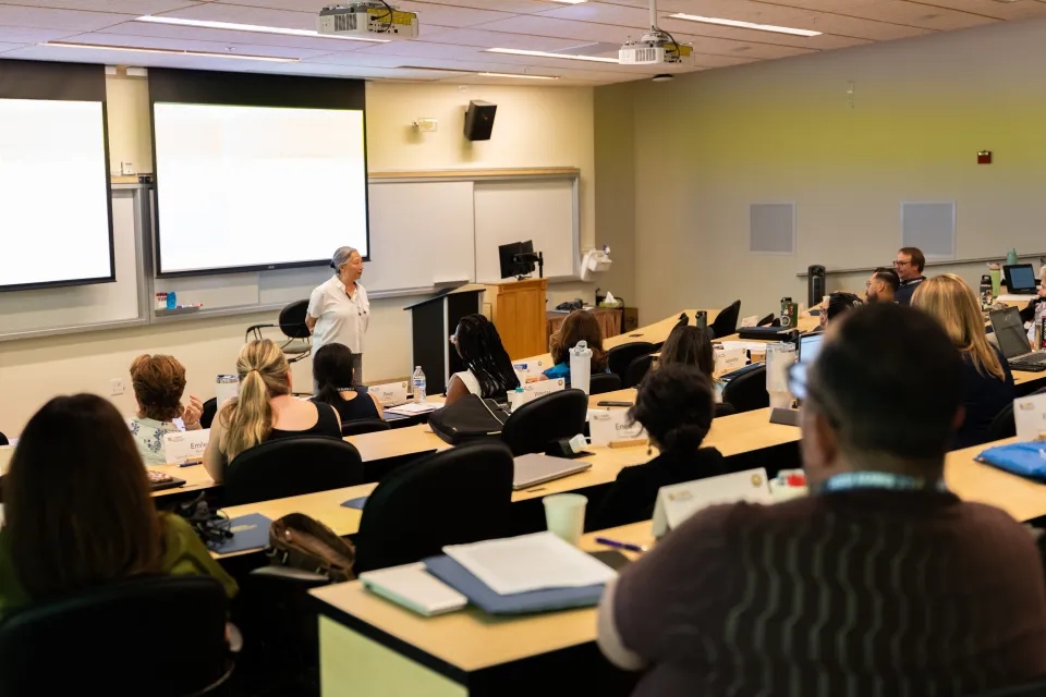 A person stands at the front of a classroom giving a presentation to an audience seated at desks, facing a screen with slides projected.