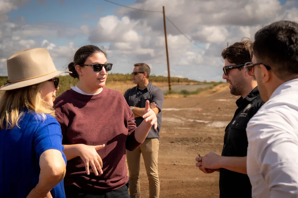 Four people stand outdoors in conversation, with another person in the background; the setting appears to be a rural or agricultural area under a partly cloudy sky.