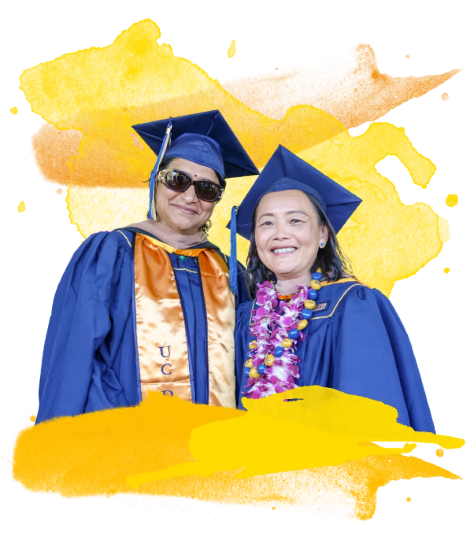 Two people in blue UC Davis graduation gowns and caps pose together; one wears sunglasses and a sash, the other has a flower lei. Yellow watercolor pattern in the background.