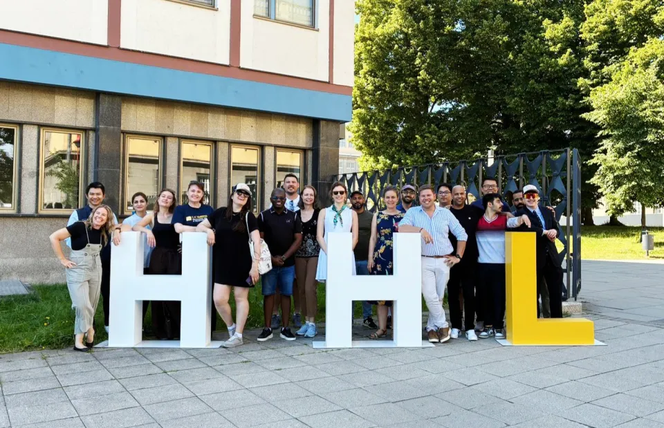 A group of people pose outdoors, smiling and standing behind large white and yellow letters spelling "HHL" on a paved area near a building and trees.