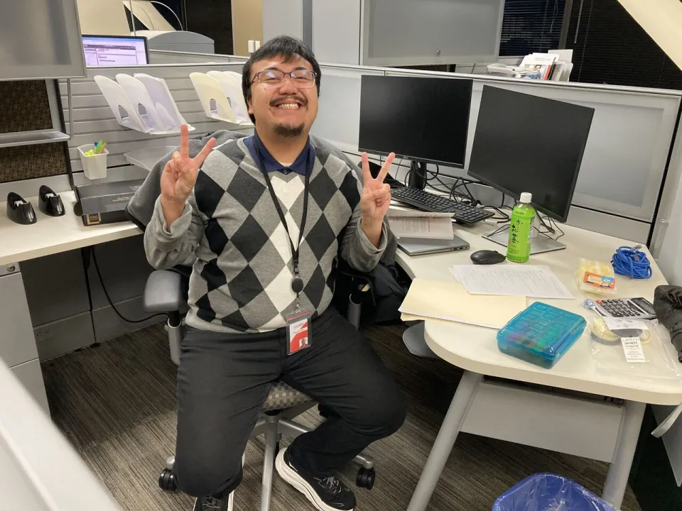 Man sitting at an office desk, smiling and making peace signs with both hands. Multiple monitors, papers, and office supplies are visible around him.