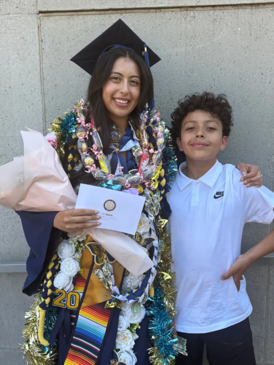 A graduate wearing a cap and gown with leis and holding flowers and a certificate stands next to a smiling boy in a white Nike shirt against a concrete wall.