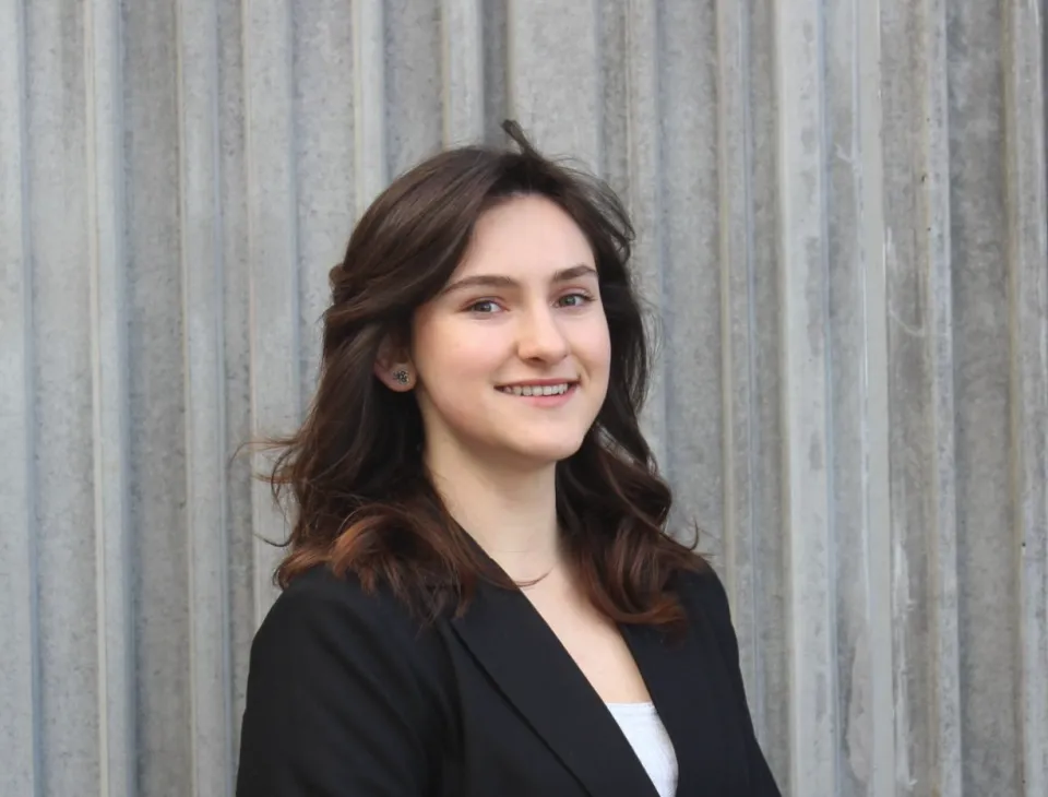 Alysa Zarkhin wearing a black blazer and white top stands in front of a corrugated metal wall, smiling slightly.