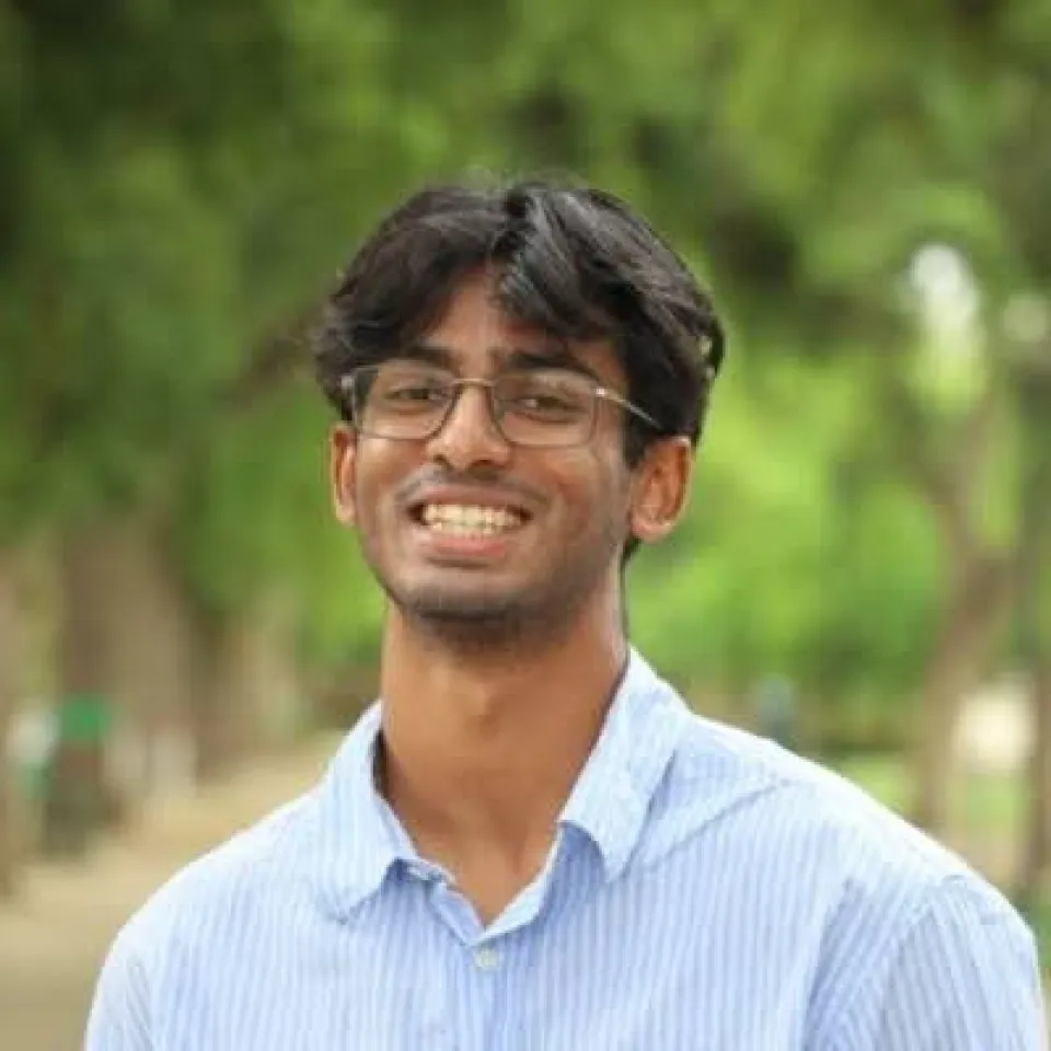 Ayan Mani smiles outdoors, with green trees blurred in the background.