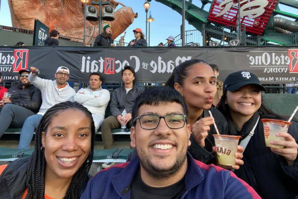 Four people in the foreground smile for a selfie at a baseball stadium, with drinks in hand. Other spectators sit in the stands behind them.