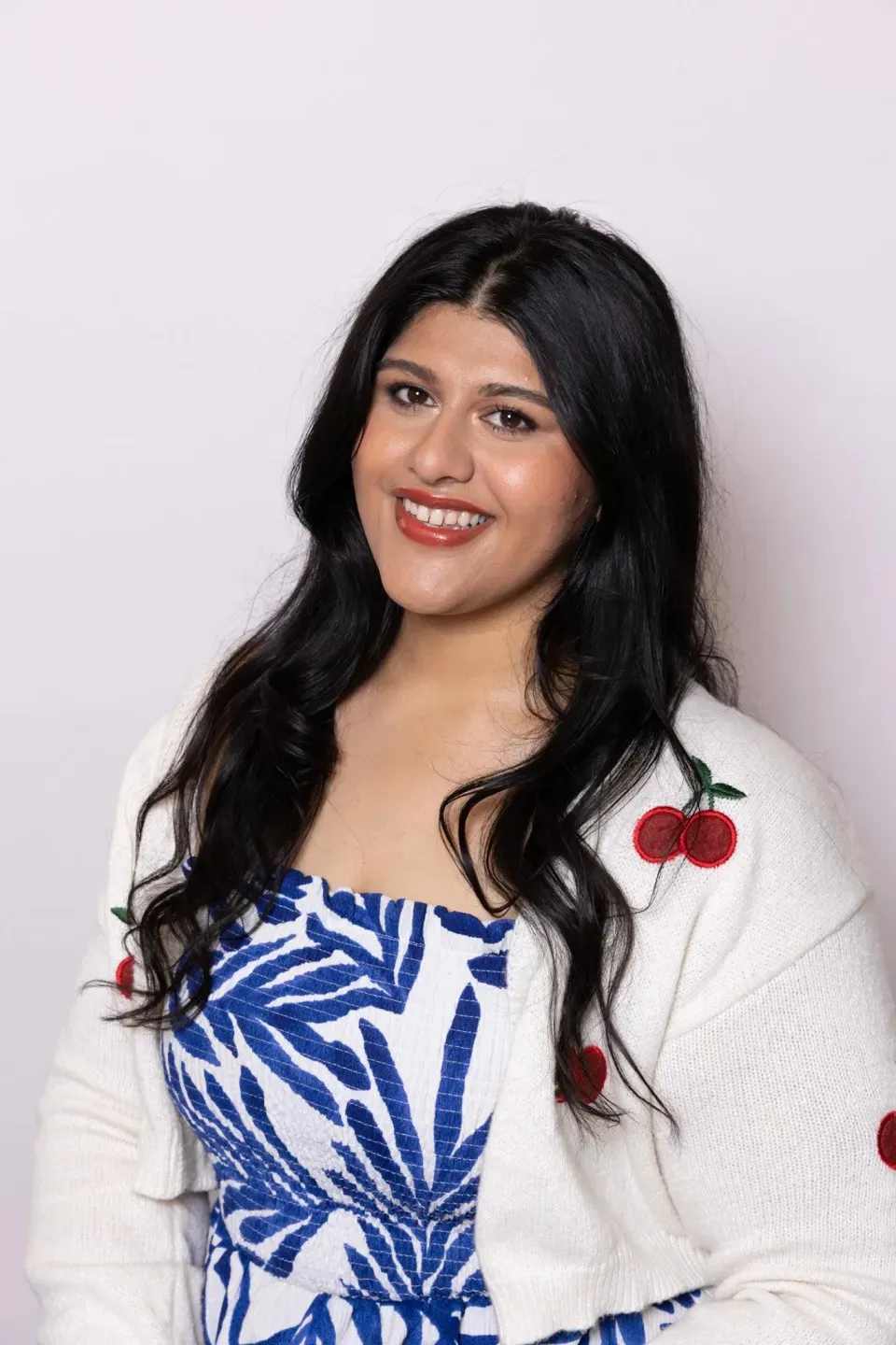 A woman with long dark hair smiles at the camera, wearing a white cardigan with cherry designs over a blue and white patterned dress, against a plain light background.