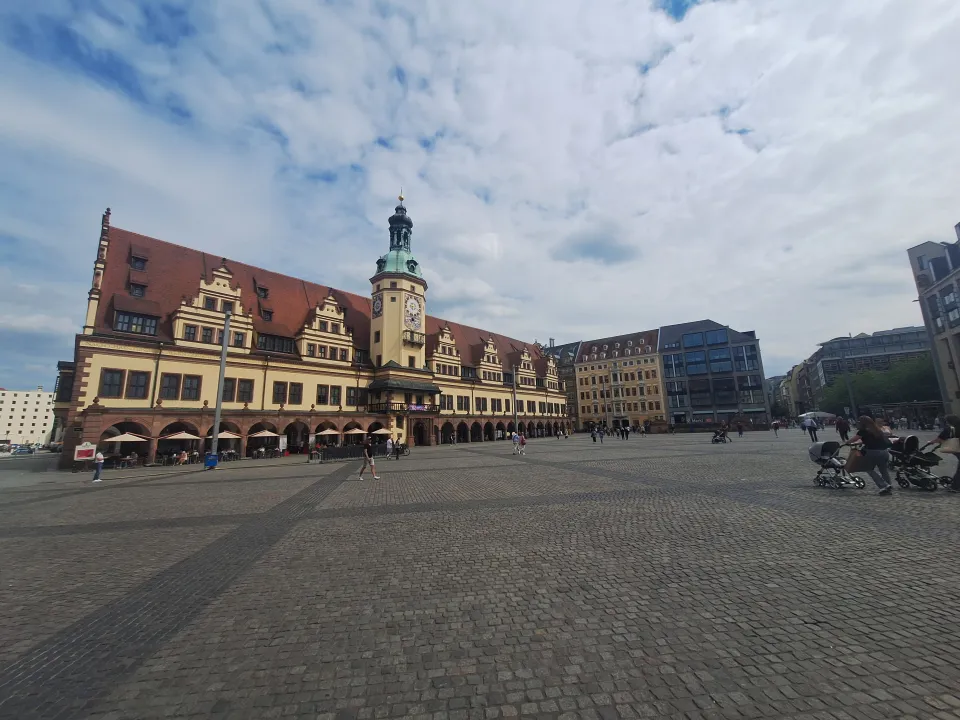 Large cobblestone square with a historic building featuring a clock tower on the left, modern buildings on the right, and people walking and sitting around.
