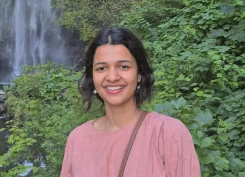 Woman in a pink top standing outdoors in front of dense green foliage and a waterfall, smiling at the camera.