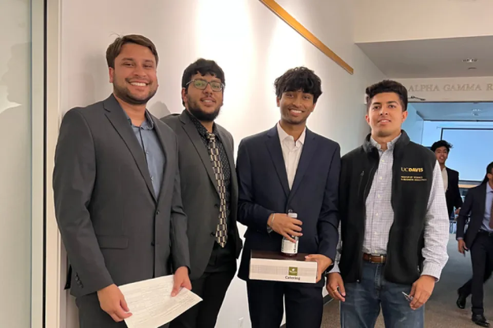 Four men in business attire stand together indoors; one holds a certificate, another holds a box and bottle. A UC Davis vest logo is visible on one person.