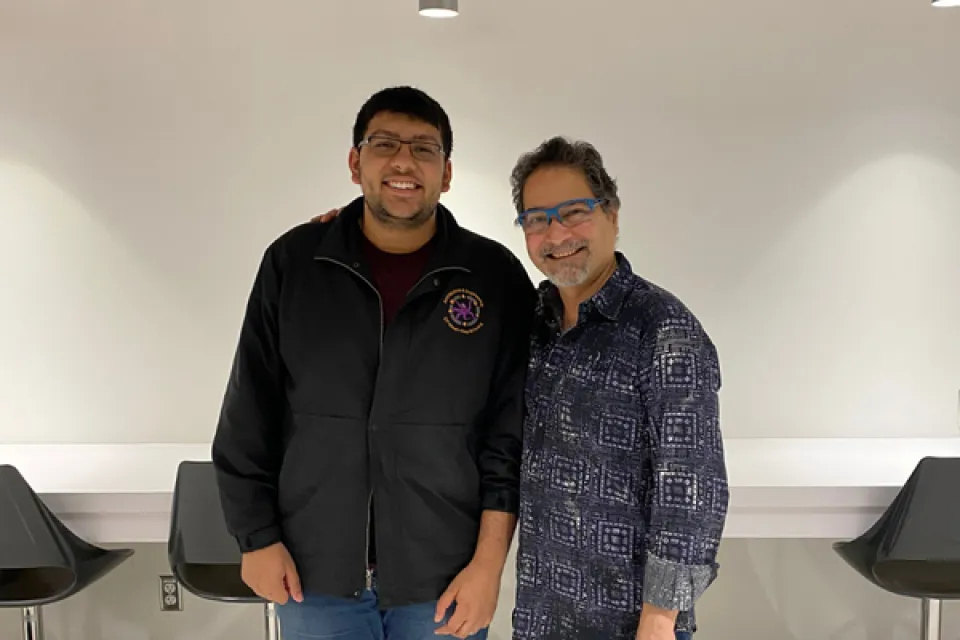 Two men are standing indoors, smiling at the camera. One has his arm around the other. They are in front of a white wall and empty black chairs.